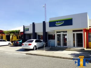 Edificio bancario de una planta con fachada moderna, cartel azul de BanReservas, tres coches aparcados y rampa accesible en la entrada.