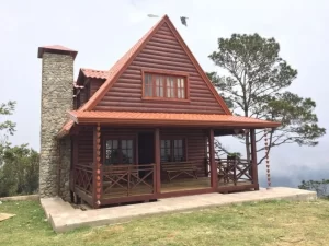Cabaña rústica de madera con techo inclinado y chimenea de piedra, rodeada de naturaleza.