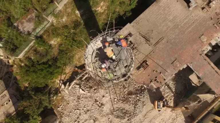 Vista aérea de cuatro obreros con equipo de seguridad trabajando sobre estructura circular entre escombros y vegetación.